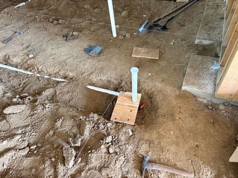 Dirt floor marked with taut string lines, a capped white PVC stub-up braced upright on a plywood scrap, with a hammer and shovel resting nearby.