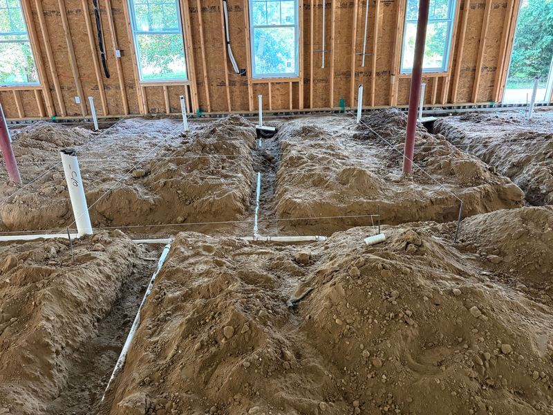 Wide view of open trenches cut across a dirt floor with white PVC drain lines laid in them, string lines stretched overhead, and framed exterior walls with windows behind.