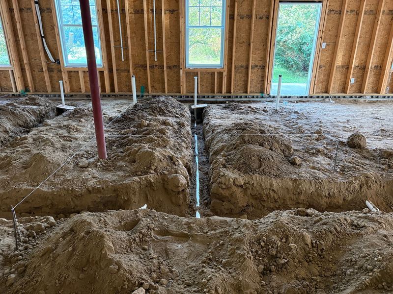 A deep trench running toward the camera with a white PVC drain line at the bottom, red steel support column to the left, and a framed wall with windows and an exterior door behind.