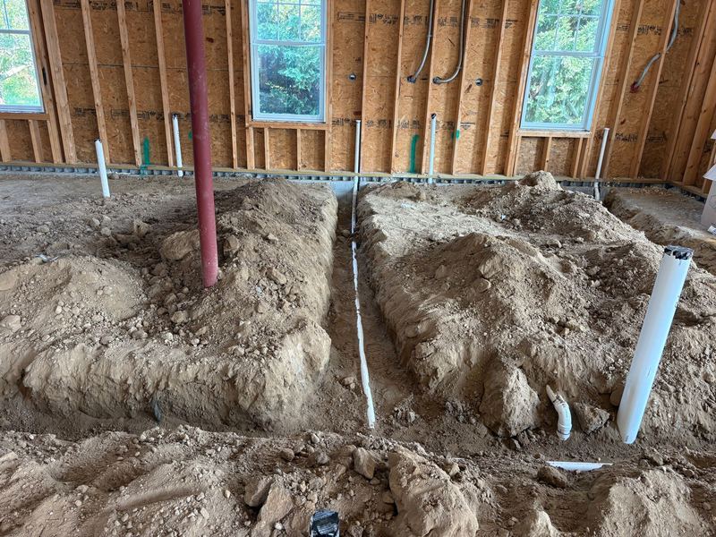 Trench running parallel to a framed exterior wall with a white PVC drain line in the base, capped stub-ups standing along the wall, and flexible electrical conduit above.