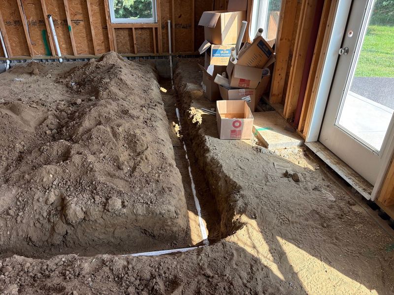 A long trench with a drain line running toward a glass exterior door, with cartons of pipe fittings stacked in the corner and daylight coming through the doorway.
