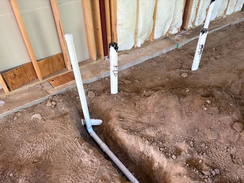 Close-up of three capped PVC stub-ups rising from a dirt floor, hand-labelled in marker, next to a trench with a drain line and a wall filled with spray foam insulation.