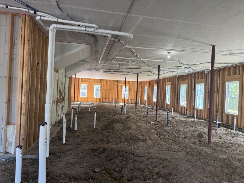 Long interior view over a backfilled dirt floor dotted with capped stub-ups, with white PVC waste lines and blue water lines running along the ceiling and red steel columns down the middle.