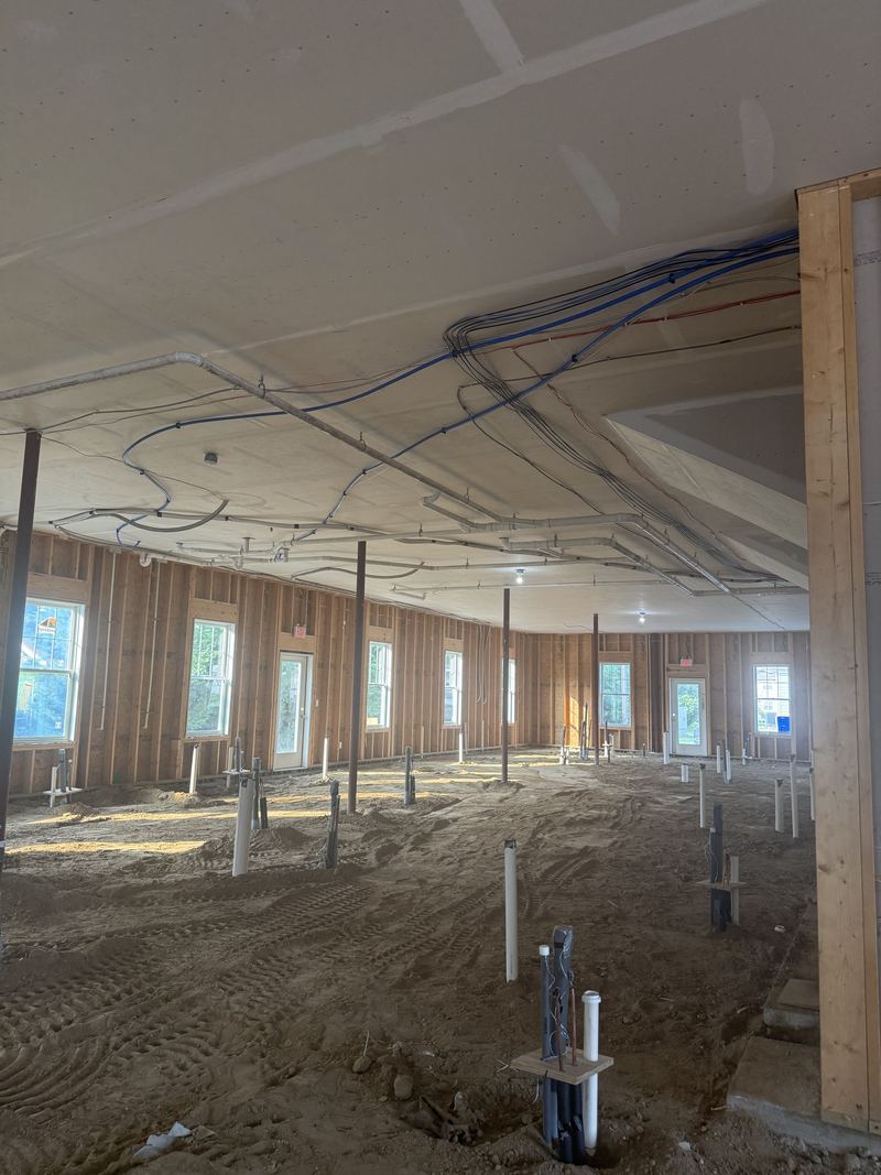 Interior with a graded dirt floor and dozens of plumbing stub-ups, looking up at a ceiling carrying white PVC pipe, blue water lines, and bundled electrical cable.