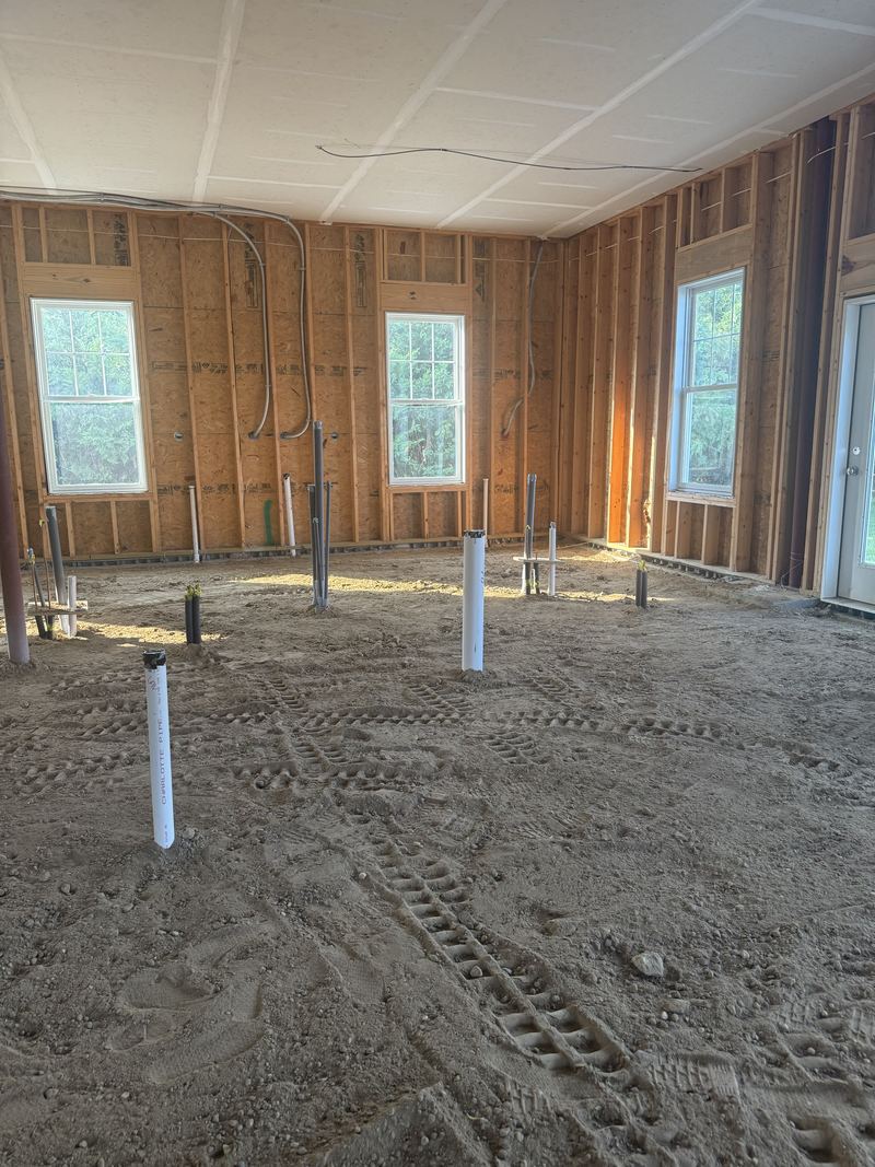 Compacted dirt floor showing roller and boot tracks, with capped white and grey stub-ups spaced across it and framed walls with windows and an exterior door behind.