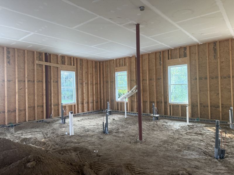 A smaller framed room with a graded dirt floor, plumbing and electrical stub-ups along the walls, a red steel column in the centre, and three windows looking onto greenery.