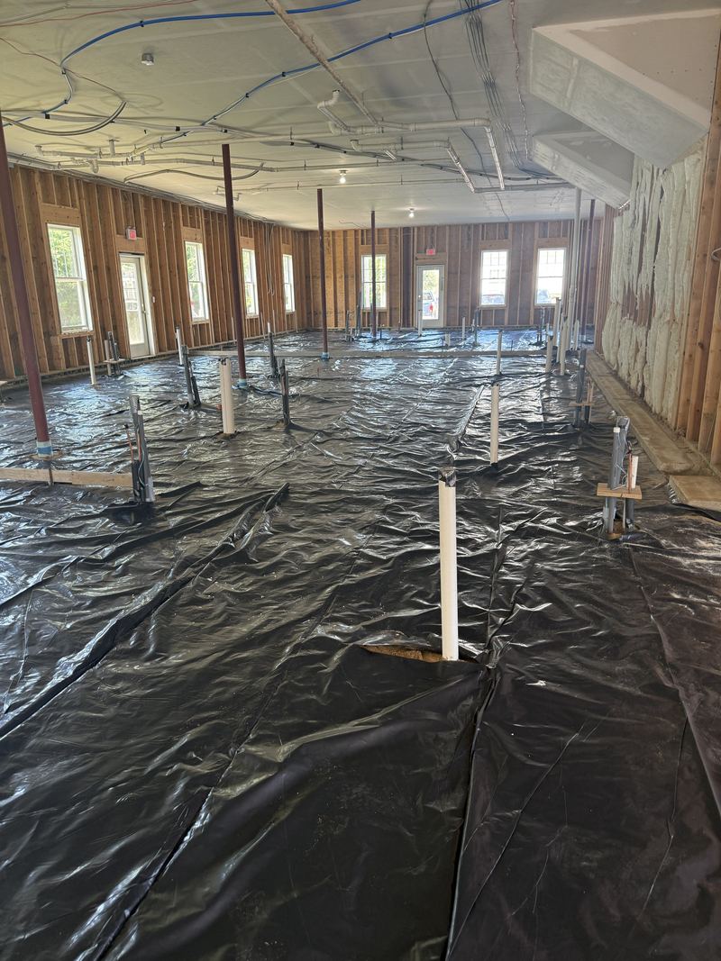 Black vapor barrier laid across the full floor with plumbing stub-ups sleeved through it, red steel columns, spray foam insulation on the right-hand wall, and overhead pipework.