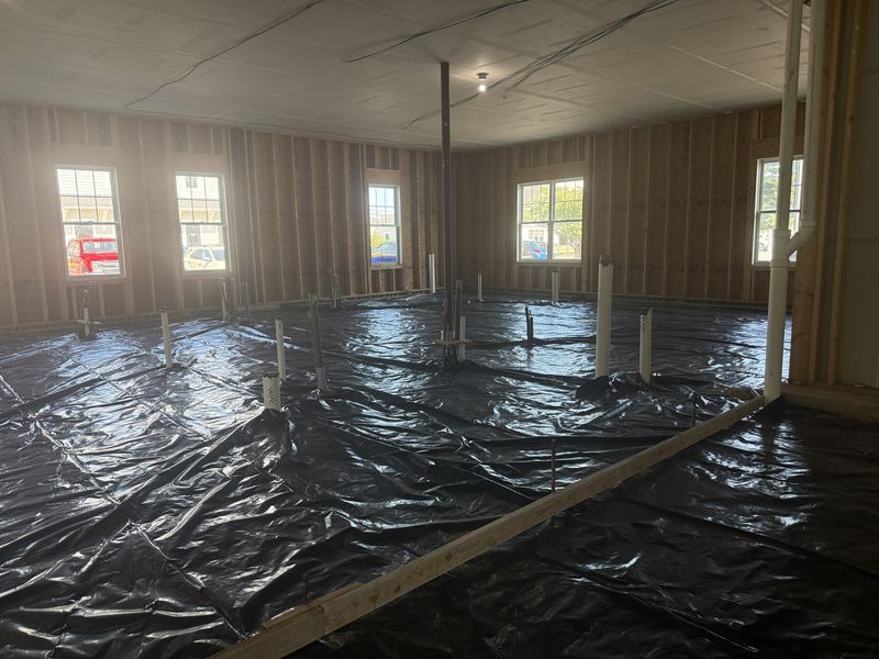Vapor barrier covering the floor with labelled stub-ups standing through it, a timber screed board laid across the foreground, and windows looking out to parked vehicles.