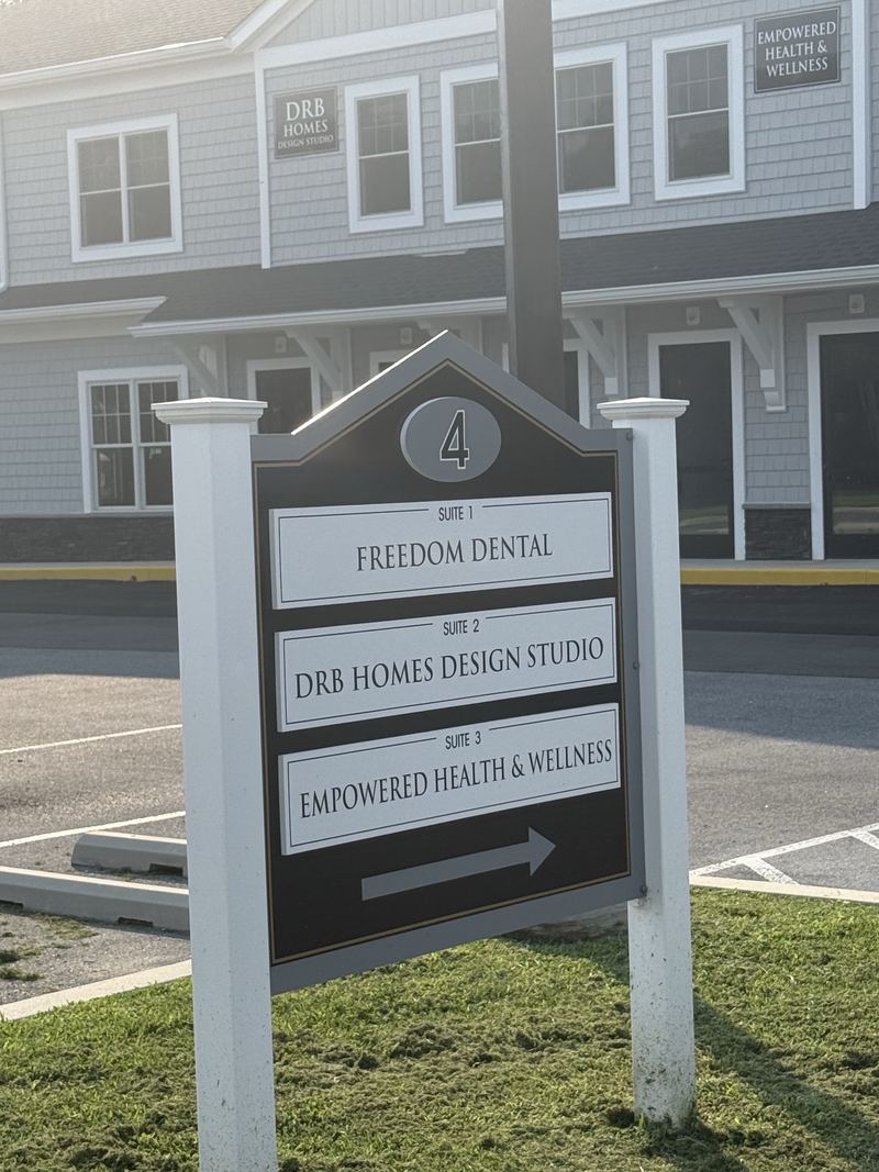 Building directory sign for building 4 at Plantations Professional Park listing three suites, with the grey shingled building behind it.